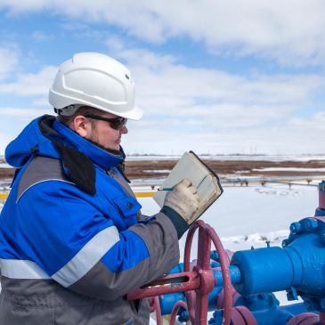 Worker in safety gear inspecting and recording data at an industrial valve outdoors.