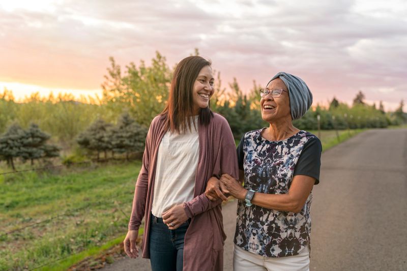 A senior woman with cancer walks with her adult daughter at sunset down a rural road. They are relaxing and staying active together. The affectionate pair are talking and walking with arms linked.