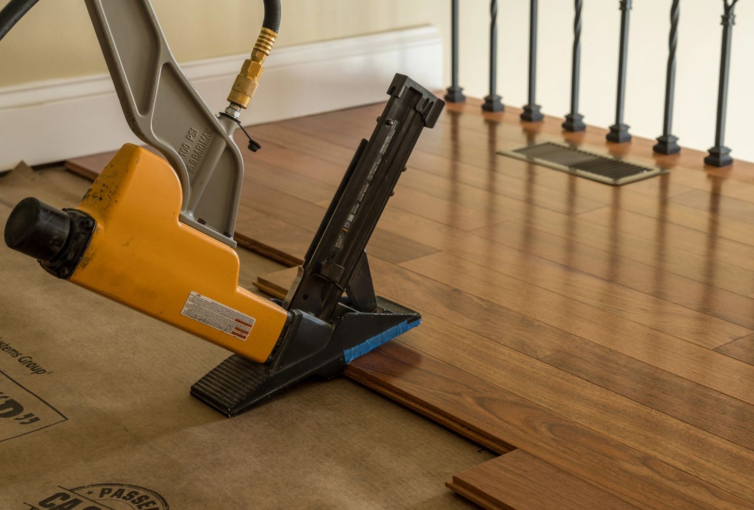 A yellow nail gun installing wooden flooring over a protective underlayer.
