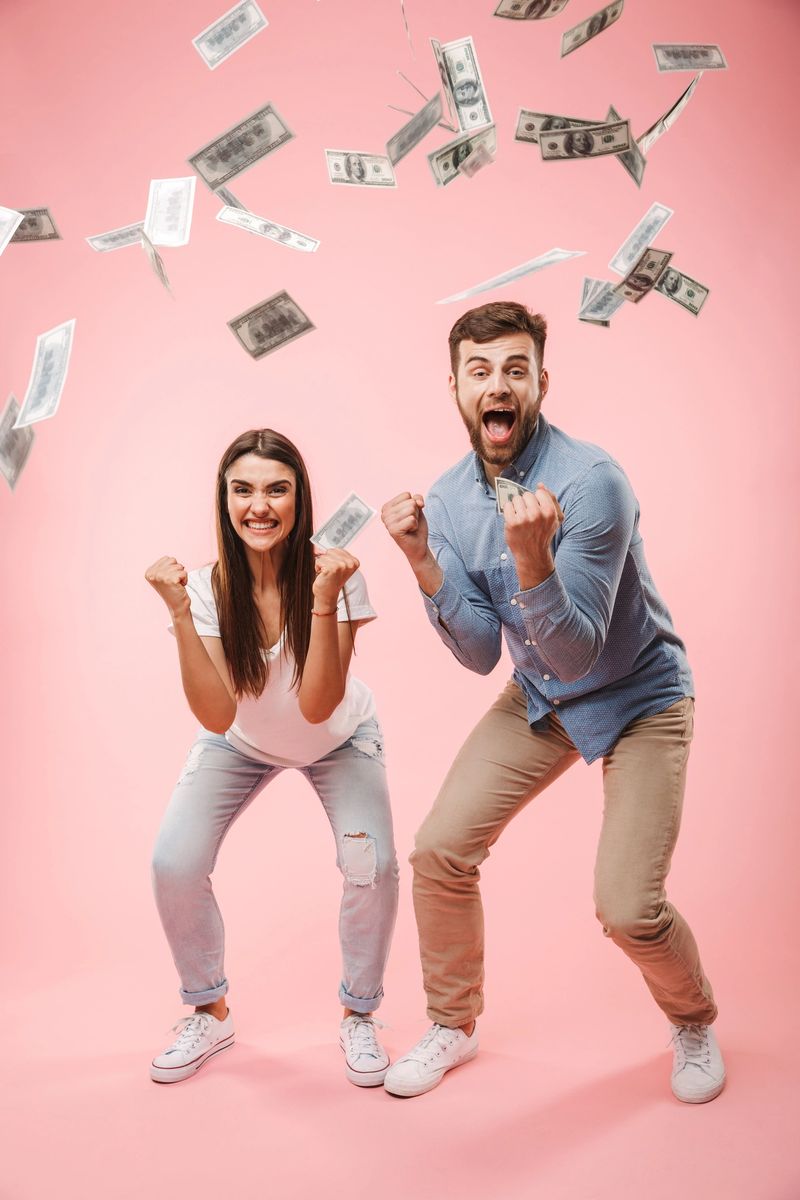 Full length portrait of an excited young couple standing under money banknotes shower and celebrating success isolated over pink background