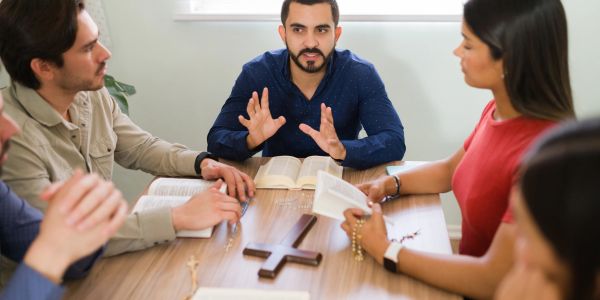 Group praying with rosaries and open Bibles around a wooden cross.