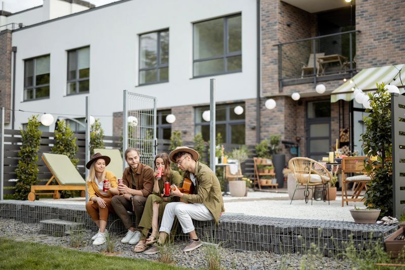 Group of young friends spending summertime together, having fun sitting together with drinks and guitar on a porch of the country house, wide view on a beautiful cottages