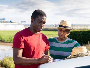 dos hombres uno de ellos anotando en un papel y el otro observando, al fondo un campo