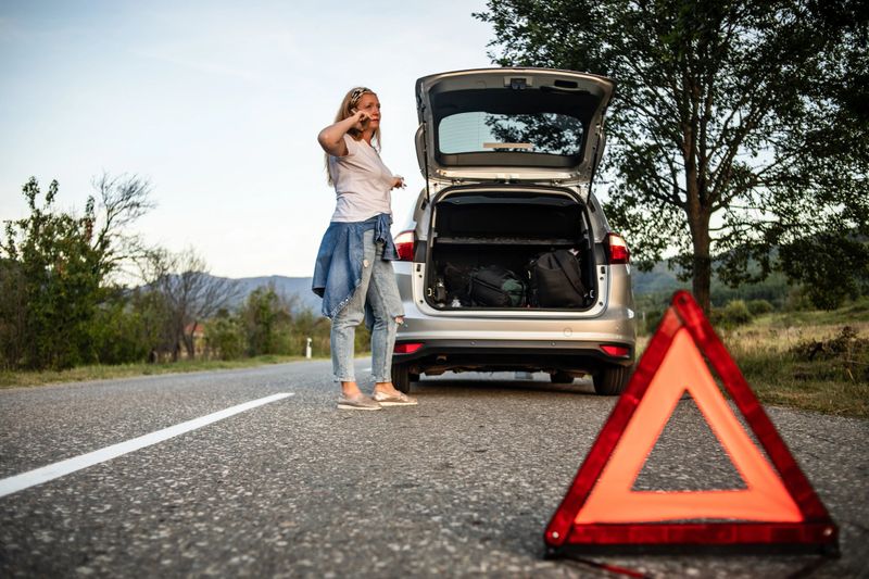 One woman is standing alone on the road next to her car and calling the roadside assistance service.