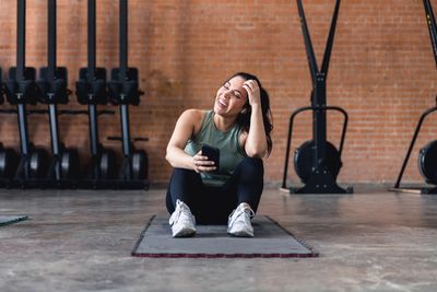 Woman laughing and holding phone while resting on gym mat.