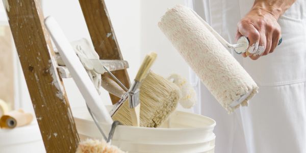 Hand holding paint roller near paint bucket and ladder.