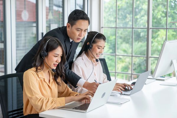A professional man assists two women wearing headsets at their laptops in a bright office.