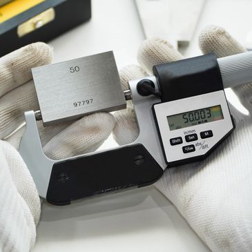 Gloved hands holding a digital micrometer measuring a metal block.