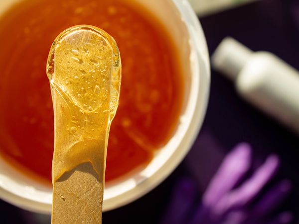 Close-up of a wooden spatula with honey-like wax over a wax pot for hair removal.