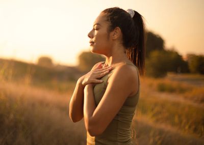 Woman practices mindfulness outdoors at sunset, hands on chest, eyes closed.
