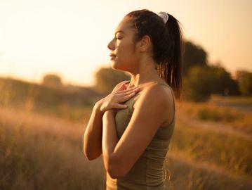 Woman practices mindfulness outdoors at sunset, hands on chest, eyes closed.