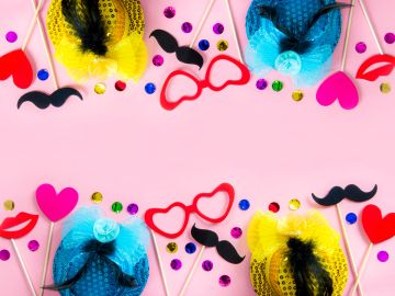 Colorful party props including hats, glasses, and mustaches arranged on pink background.