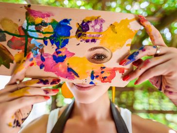 A woman with paint on her hands holds a colorful palette in front of her face.
