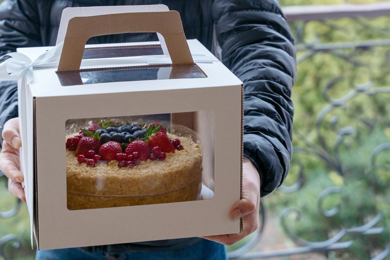 Human hands holding cardboard box with holiday cake decorated of berries. Spring time, person on the front door, courier or family member bringing the dessert. Outdoors, copy space, garden background.