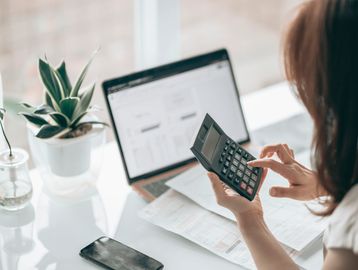 Person using a calculator with a laptop and documents on a desk.