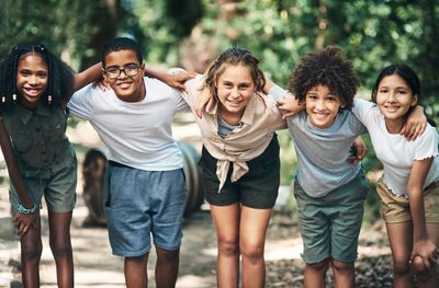 Five diverse kids smiling with arms around each other outdoors.
