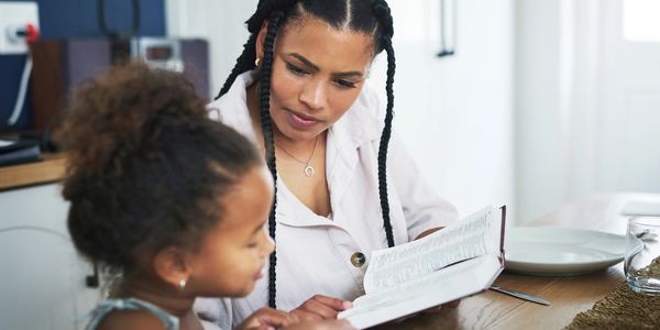 A woman reading a book to a young girl at a dining table.