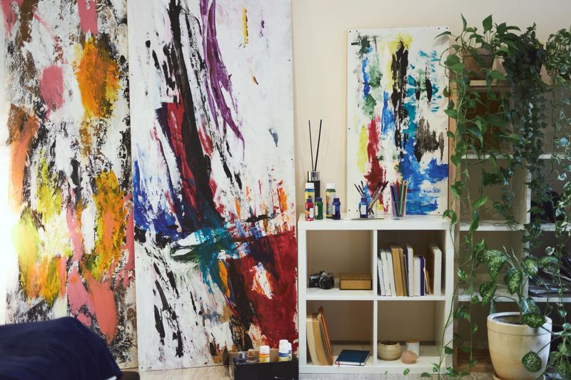 Abstract art paintings on wall of stylish modern living room interior with bookcase, home plants and paints. Home hobbies, interior ideas concept