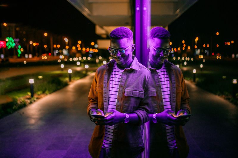 Young handsome black man using phone, typing text message in a front of neon blue light office window in rainy night