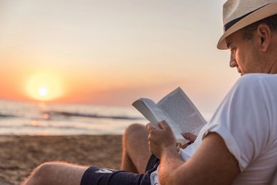 Man reading a book on the beach at sunset wearing a hat.