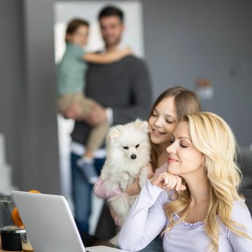 A woman working on a laptop with family and a small dog nearby.