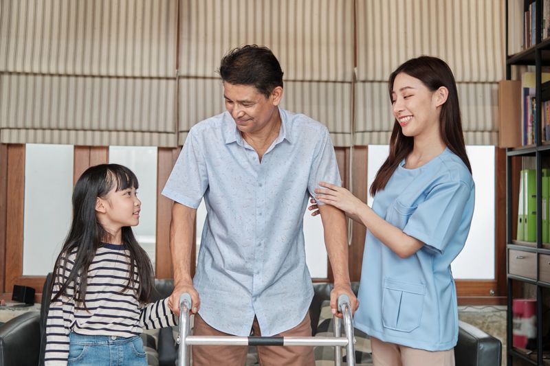 Asian female physiotherapist helping, training, and rehabilitating an elderly disabled man in a private home. Healthcare guiding, physical recovery with a granddaughter supporting beside.