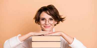 Smiling woman with glasses resting on a stack of books against peach background.