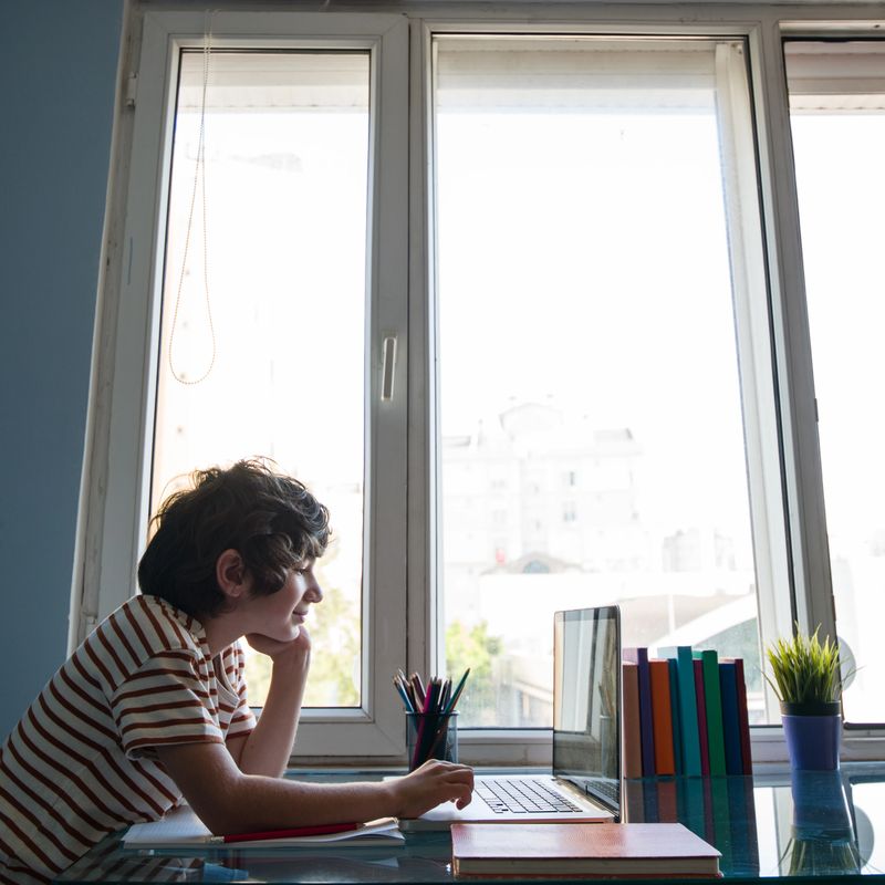 Photo of 8 years old elementary schoolboy sitting next to window and homeschooling. He is using a laptop computer for online video conferencing class. Shot indoor with a medium format camera.