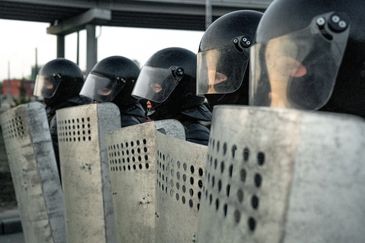 Line of riot police with helmets and shields standing in formation.