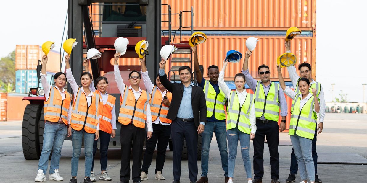 Diverse group of workers celebrating with safety helmets raised at a shipping yard.