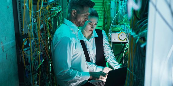 Two IT professionals working on a laptop in a server room filled with cables.