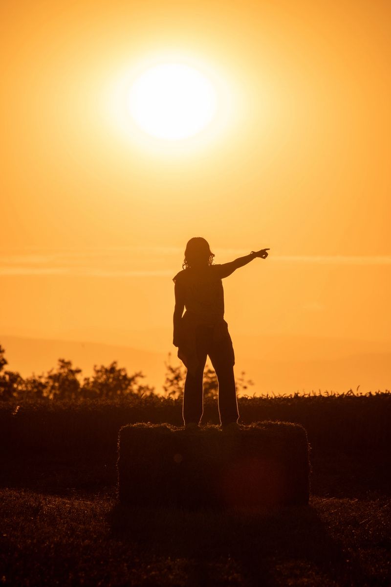 silhouette of woman standing on a bale of straw with the sun against the backlight pointing to the horizon at sunset. photography with vertical format. Pointing a finger to infinity