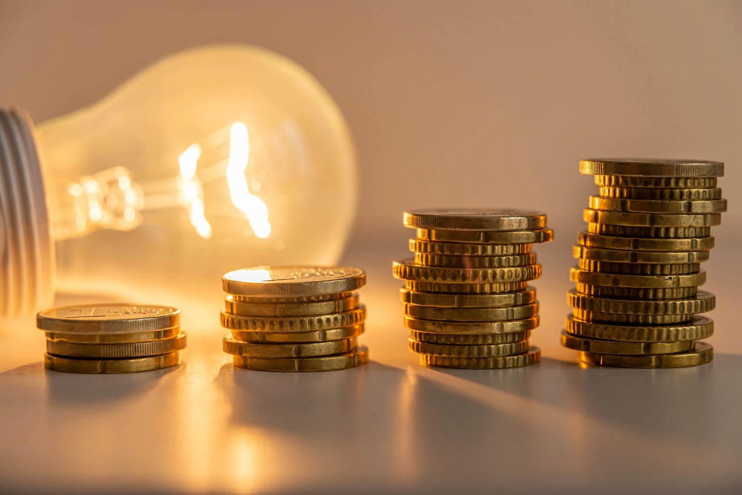 Stacks of golden coins with a glowing light bulb in the background.