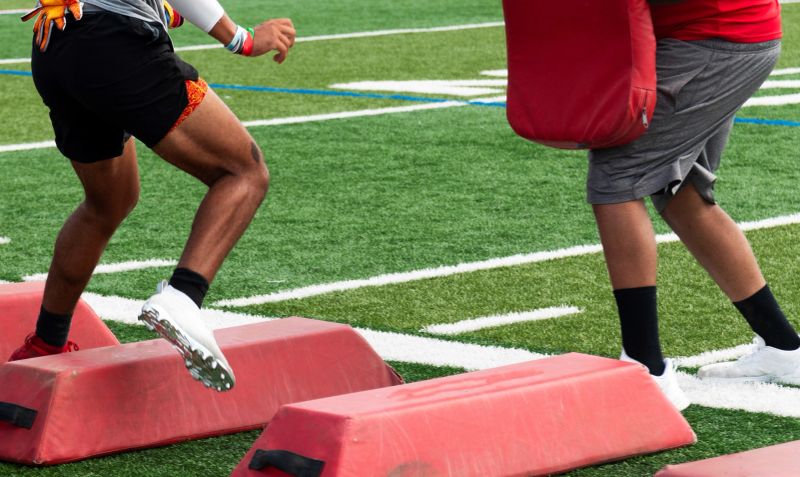 Football player running sideways over red barriers while taking a hit by the coach with a red pad at summer camp