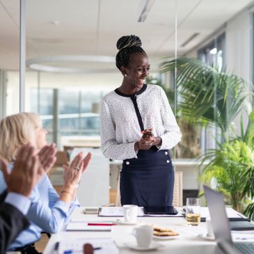Woman standing and smiling as colleagues applaud in a meeting.