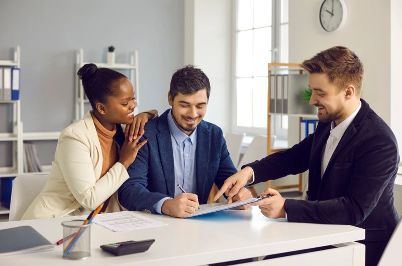 Successful meeting with broker bank agent insurer. Happy mixed ethnicity couple customers sign mortgage loan contract. Smiling african american wife hugs caucasian husband putting signature on paper