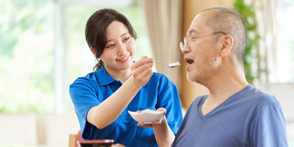 A caregiver feeds an elderly man with a spoon, smiling warmly.