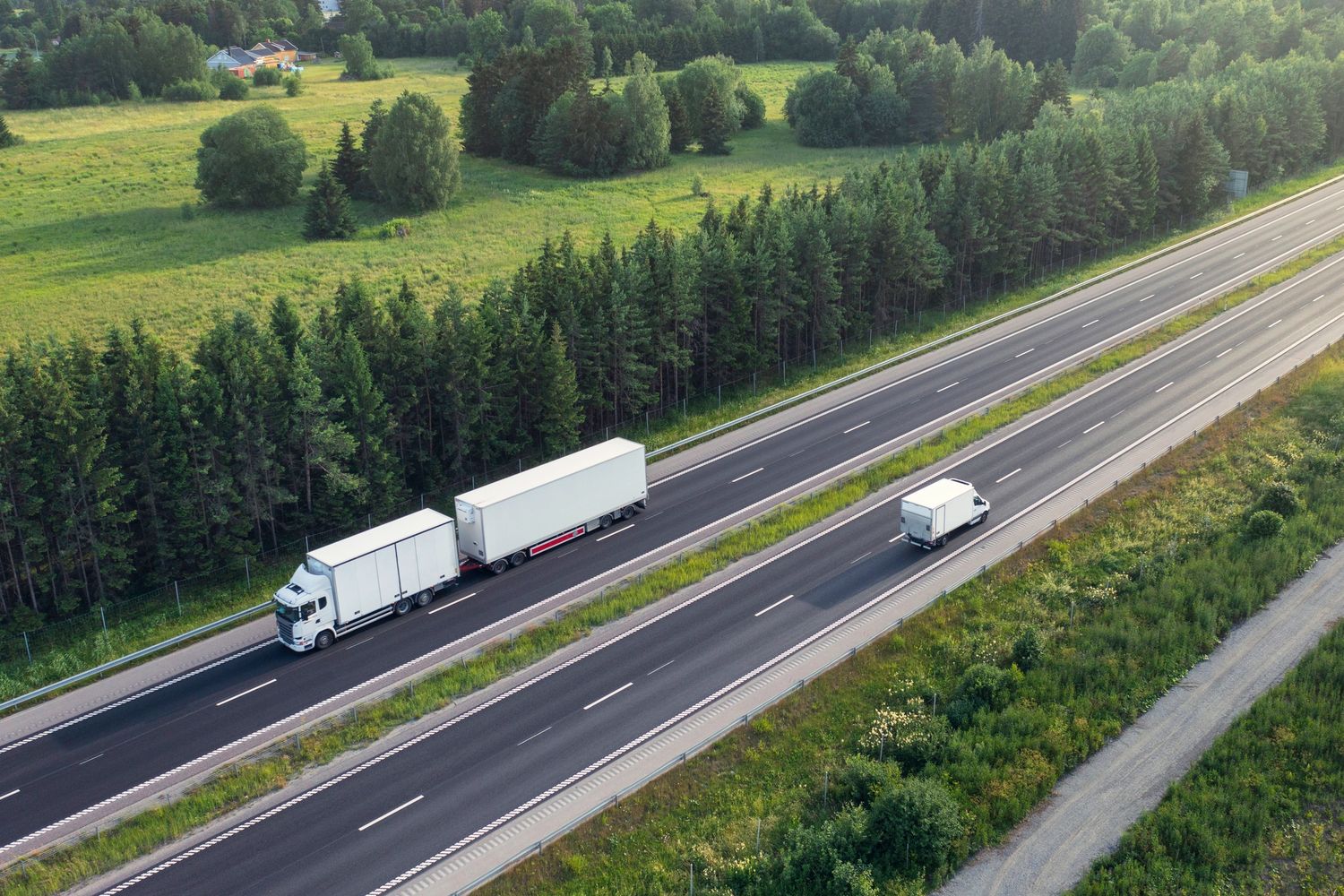 Two white trucks and a van driving on a highway surrounded by green fields and trees.