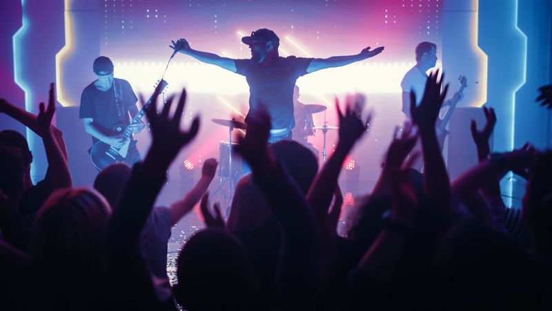 Rock Band with Guitarists and Drummer Performing at a Concert in a Night Club. Front Row Crowd is Partying. Silhouettes of Fans Raise Hands in Front of Bright Colorful Strobing Lights on Stage.