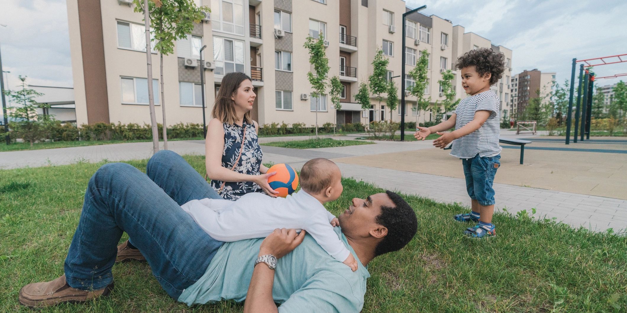 Family playing on grass in an urban park near apartment buildings.