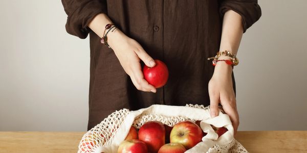 Person holding a red apple next to a net bag filled with apples on a wooden table.
