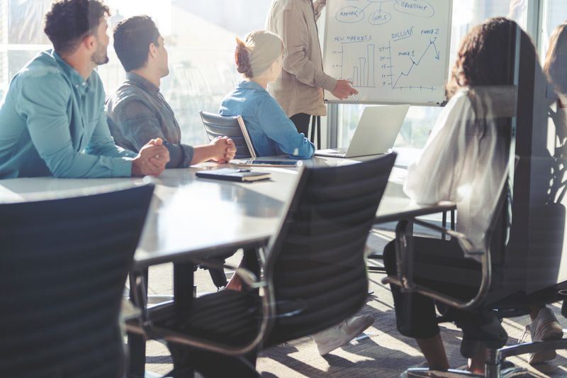 Business people watching a presentation on the whiteboard. A mature man is writing on the whiteboard with charts and graphs. They are sitting in a board room, there are laptop computers and technology on the table. All are casually dressed. There is a window behind him with city views.