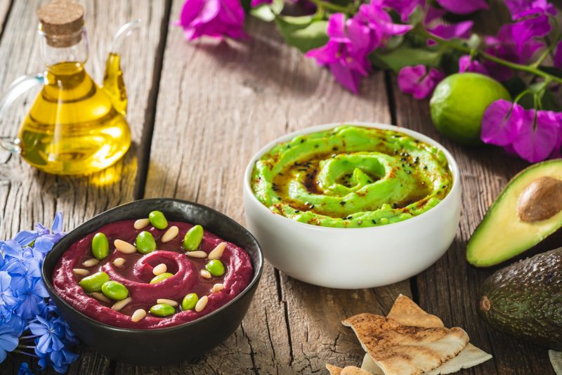 Beetroot Hummus and Avocado Hummus bowls on wood table with flowers and olive oil as Mediterranean food