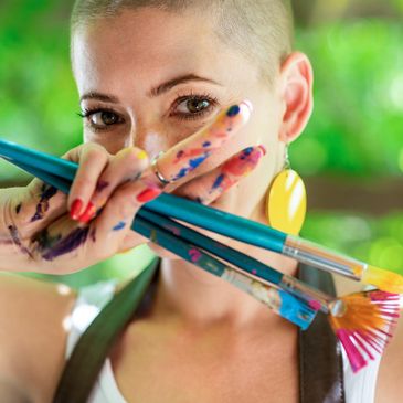 Person holding paintbrushes with paint on fingers near face.