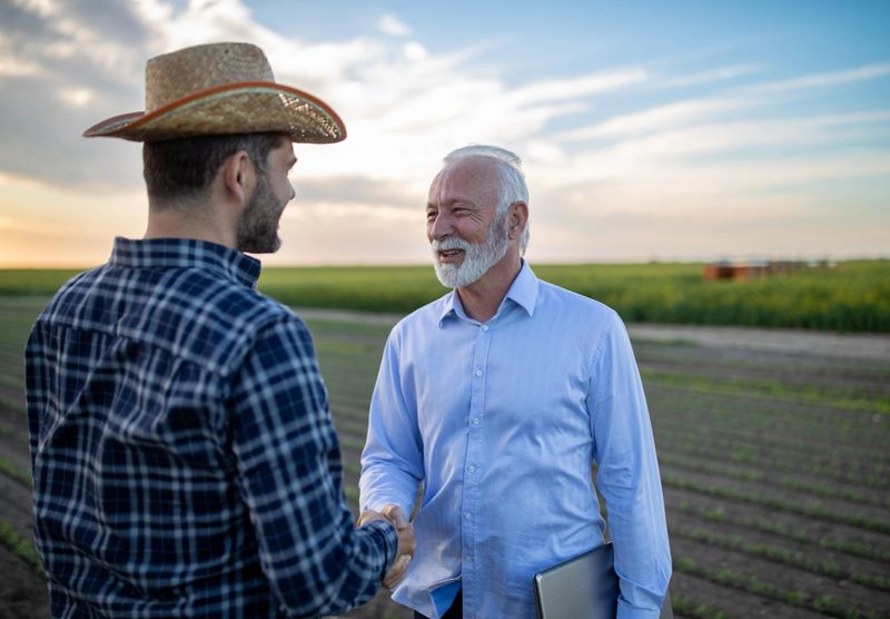 Farmer and insurance sales rep representative reaching agreement deal. Two men agronomist shaking hands standing in field happy satisfied.