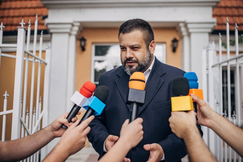 One man, male politician talking into reporters microphones outdoors.