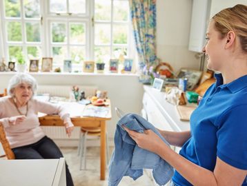 A young woman is drying a dish while talking with a senior who is sitting at the table nearby.