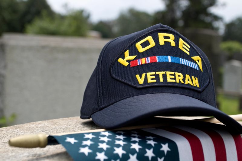 Korean War Veterans cap inscribed with text depicting the Korean War from June 1950-July 1953, sitting on tombstone with American Flag.