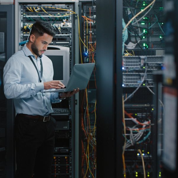 IT technician working on a laptop in a server room surrounded by network equipment.