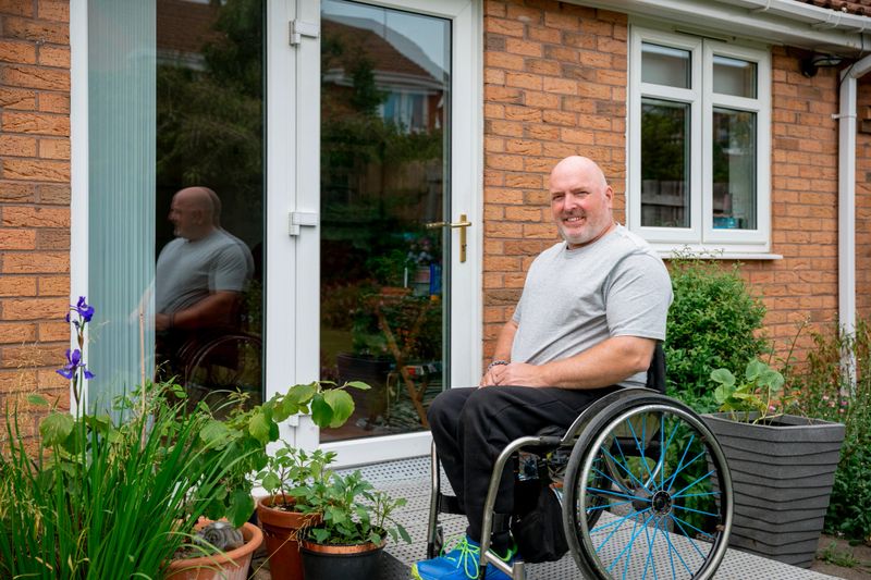 A portrait shot of a caucasian, mature man with paraplegia, outside of his home on a wheelchair ramp, looking and smiling at the camera.